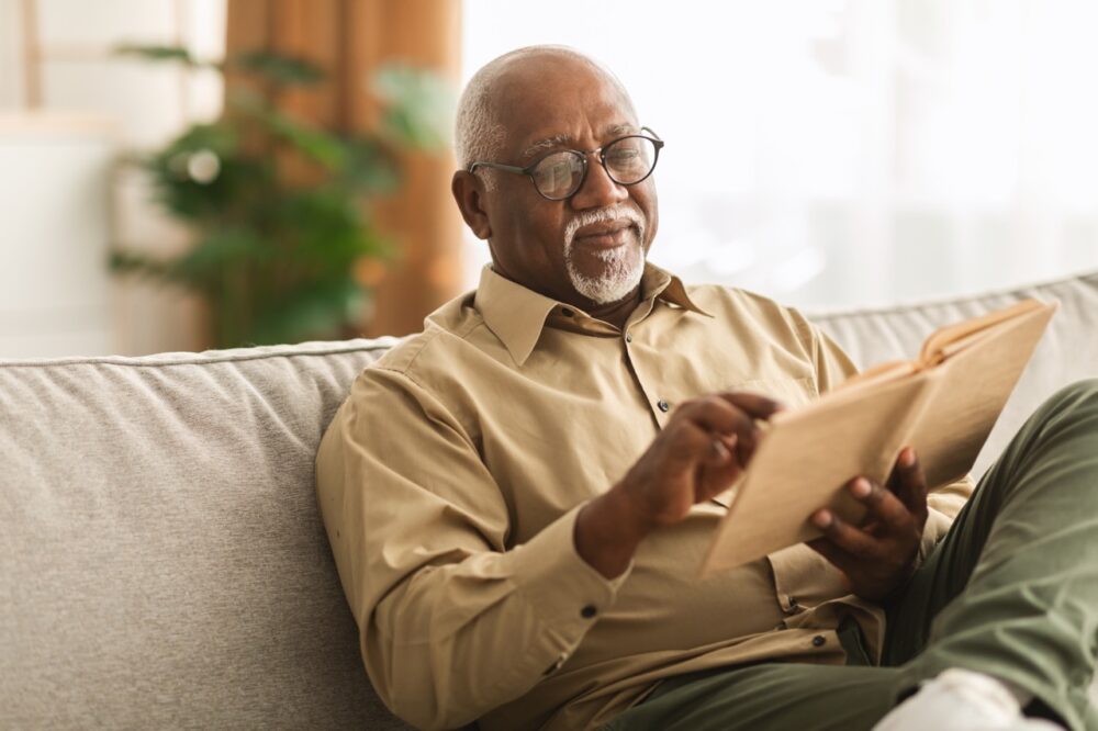 Senior,African,American,Man,Reading,Book,Sitting,On,Couch to show what life would be like living in a new later living home in stevenage bu Anchor New Homes