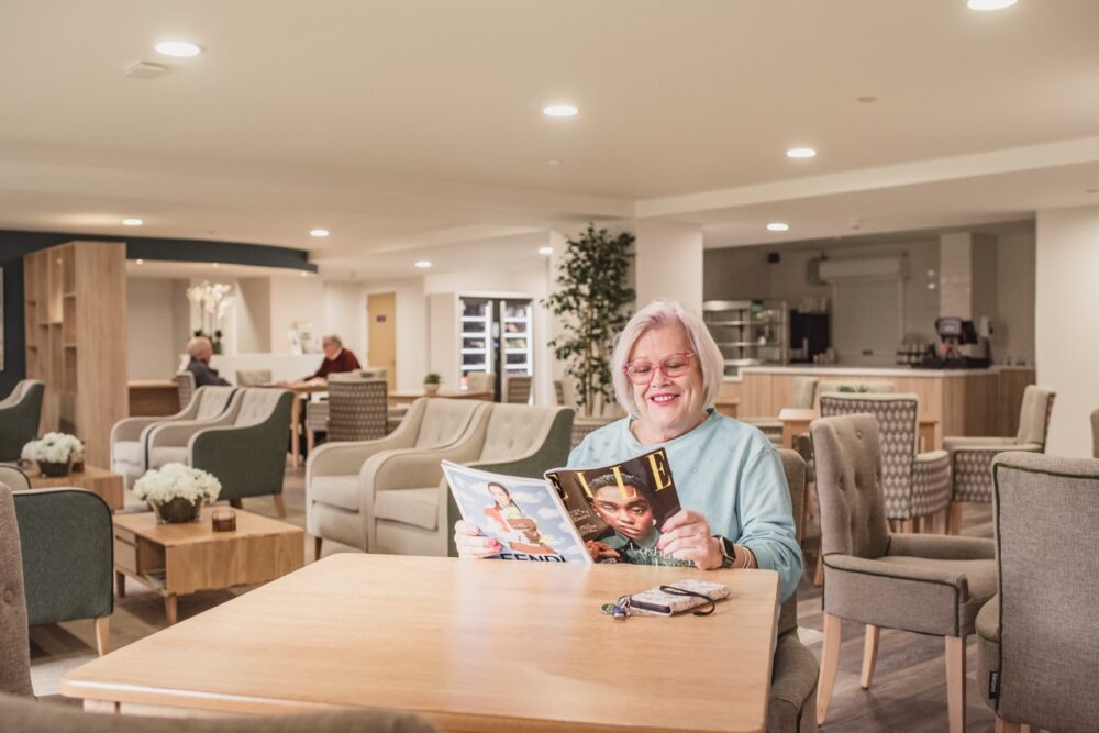 Older lady in The Dials Communal lounge readying an Elle magazine while her neighbours talk in the background. The room is neutrally decorated to a luxury standard.